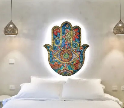 Bright boho bedroom with crisp white bed, glowing colorful Hamsa hand wall art above the headboard, woven pendant lights and minimalist side tables.