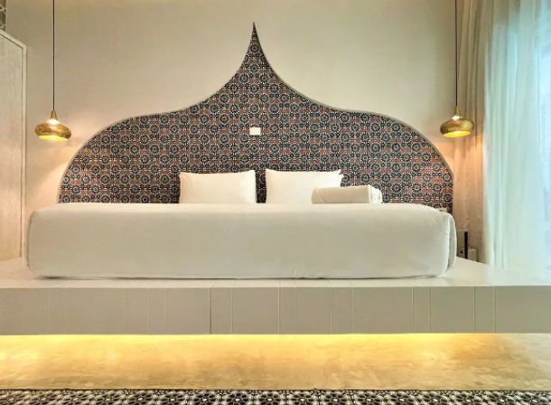 Boutique hotel bedroom interior with a raised platform king bed, crisp white linens, Moroccan-inspired arched patterned headboard, brass pendant lights, sheer curtains and warm underlighting.
