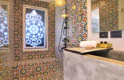 Colorful Moroccan-style mosaic bathroom with floral starburst patterned tiles on walls and bench, frosted window casting decorative light, black handheld shower, concrete sink countertop with folded white towel and toiletries
