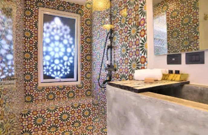 Colorful Moroccan-style mosaic bathroom with floral starburst patterned tiles on walls and bench, frosted window casting decorative light, black handheld shower, concrete sink countertop with folded white towel and toiletries