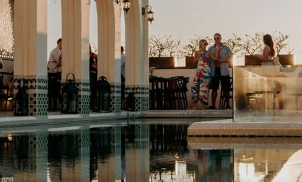Rooftop pool terrace at sunset with tiled arches and lanterns, a small group socializing — a woman in a colorful dress and companions — reflected in the calm pool water.