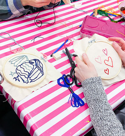 Hands working on embroidery hoops at a pink-and-white striped craft table — red stitched hearts and a blue stitched design, scissors, needles and colorful embroidery floss for a DIY sewing workshop.