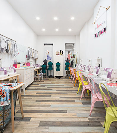 Bright sewing studio interior with rows of white sewing machines, colorful pastel metal chairs, dress forms at the back, hanging fabrics and mixed wood plank flooring