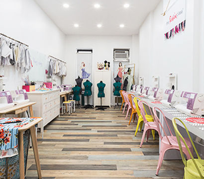 Bright sewing studio interior with rows of white sewing machines, colorful pastel metal chairs, dress forms at the back, hanging fabrics and mixed wood plank flooring