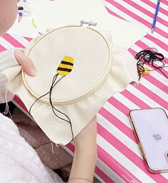 Close-up of a hand embroidery hoop with a yellow-and-black bee body being stitched, black floss trailing across cream fabric on a pink-and-white striped table, with a smartphone and threads nearby.