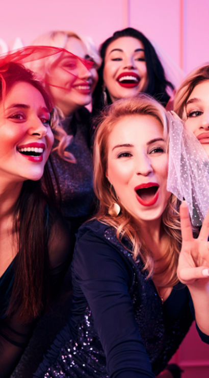Five women laughing and posing together at a bachelorette-style party, wearing small veils and making a peace sign in front of a pink backdrop with balloons.
