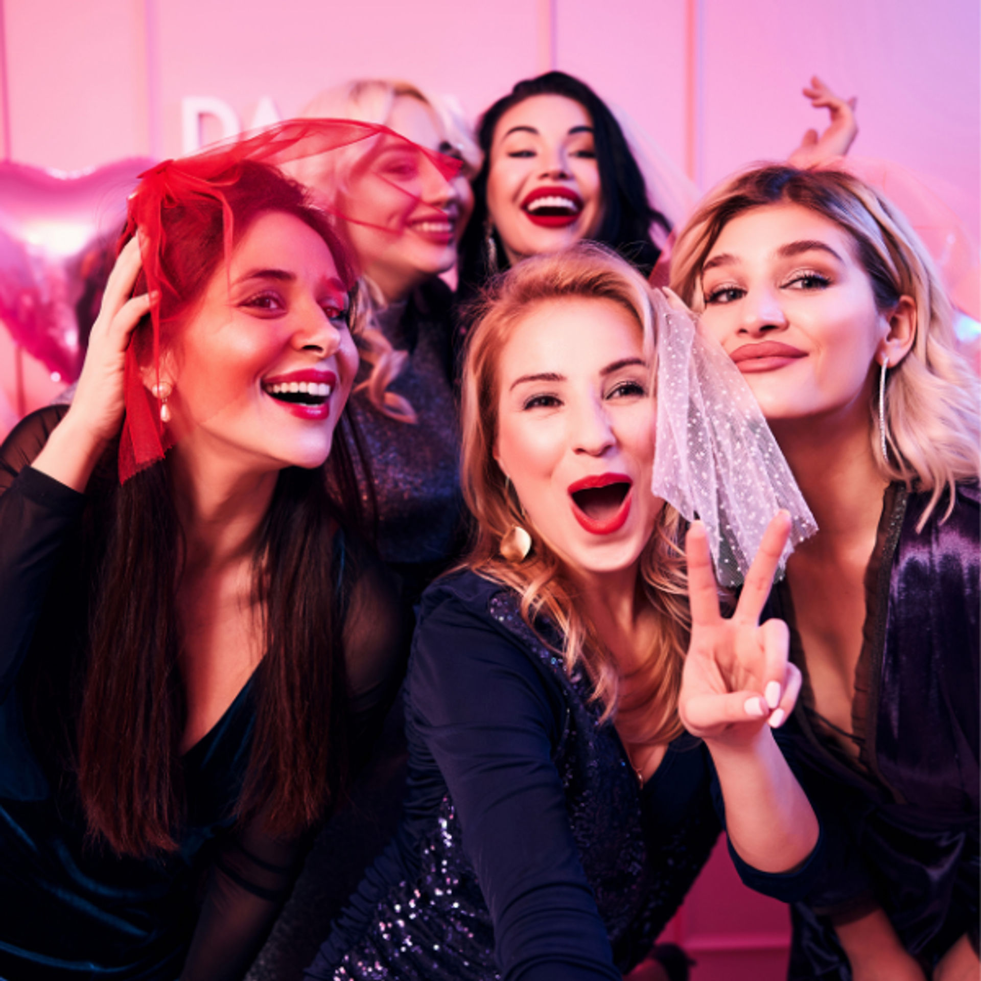 Five women laughing and posing together at a bachelorette-style party, wearing small veils and making a peace sign in front of a pink backdrop with balloons.