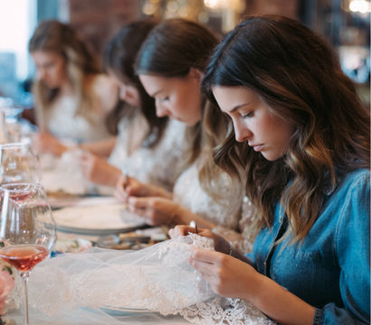 Bridal party hand-sewing delicate lace veil at an intimate indoor craft table with rosé wine and focused guests