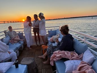 Group of friends relaxing on a cushioned boat deck during a coastal sunset cruise, chatting and sipping drinks with a bridge and calm bay waters in the background.