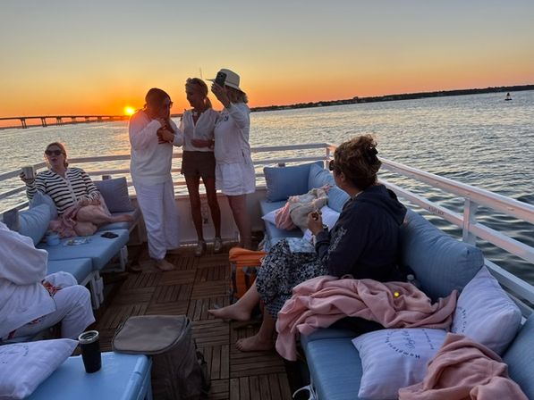 Group of friends relaxing on a cushioned boat deck during a coastal sunset cruise, chatting and sipping drinks with a bridge and calm bay waters in the background.