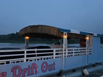 Tiki-roofed pontoon boat with a thatched canopy and glowing lanterns, docked at dusk on a calm river with a tree-lined shoreline.