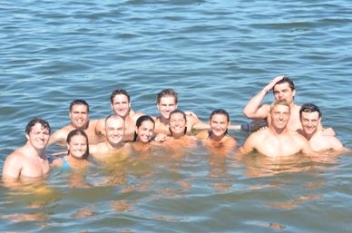 Smiling group of friends swimming together in a sunlit lake, heads and shoulders above calm blue water on a summer day