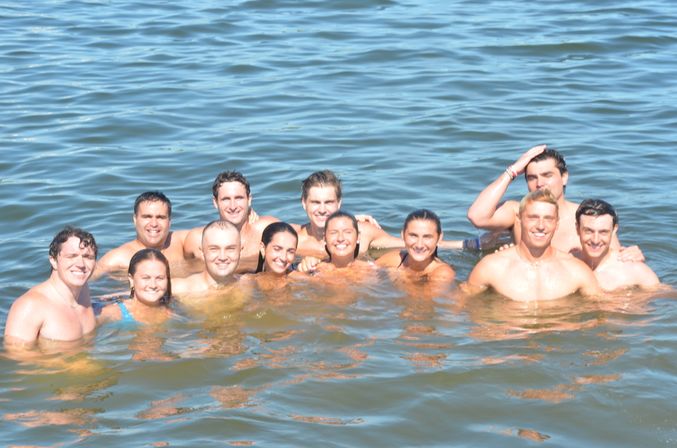 Smiling group of friends swimming together in a sunlit lake, heads and shoulders above calm blue water on a summer day