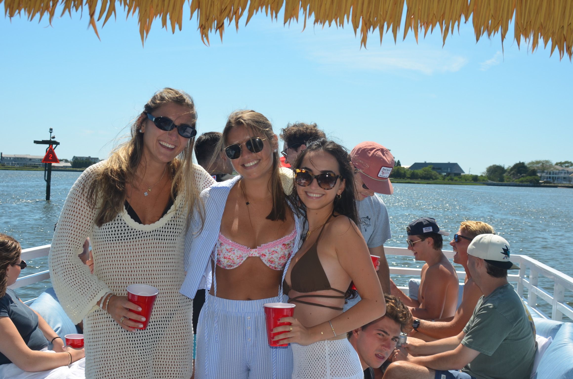 Three women in sunglasses and swimwear smiling under a thatched canopy on a sunny boat party, holding red cups while friends relax on the water with coastal homes in the background.
