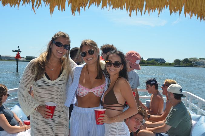 Three women in sunglasses and swimwear smiling under a thatched canopy on a sunny boat party, holding red cups while friends relax on the water with coastal homes in the background.