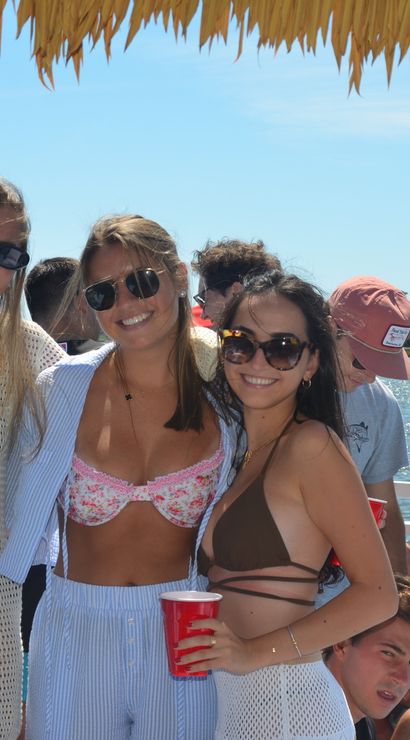 Three women in sunglasses and swimwear smiling under a thatched canopy on a sunny boat party, holding red cups while friends relax on the water with coastal homes in the background.