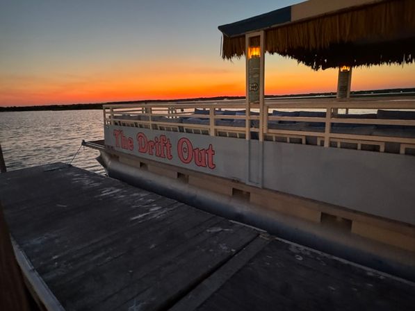 Pontoon boat moored at a wooden dock during a vibrant lake sunset, calm water reflecting orange and purple sky, thatched tiki canopy and glowing lantern.