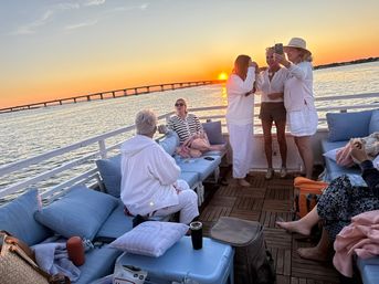 Coastal sunset boat cruise with friends lounging on cushioned deck seating, woman taking a selfie, bridge spanning calm water