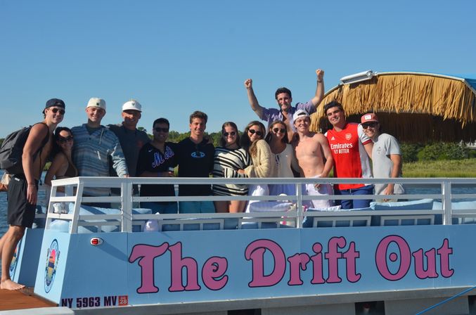 Group of young adults posing on a blue party pontoon with a tiki-style canopy at a sunny dock, smiling and celebrating over calm marsh waters