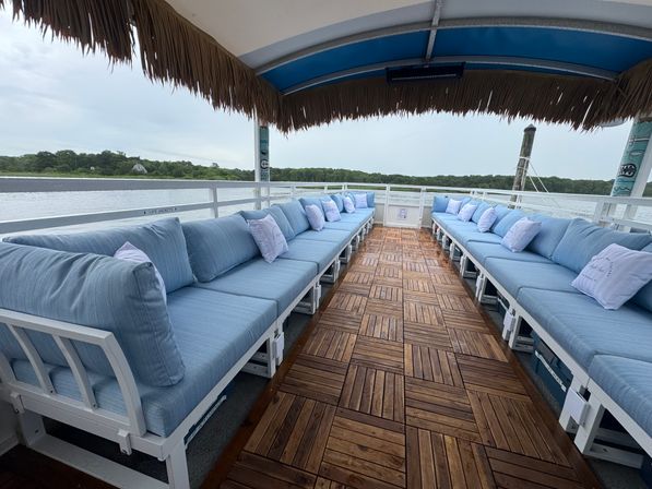 Spacious waterfront pontoon lounge with twin rows of light-blue cushioned benches, teak-style wooden deck tiles and a thatched tiki canopy, overlooking a calm river and tree-lined shoreline.