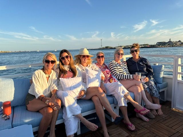 Six friends lounging on a coastal boat deck at golden hour, seated on a cushioned bench with calm harbor water and shoreline buildings in the background — casual summer outfits, sunglasses and drinks.