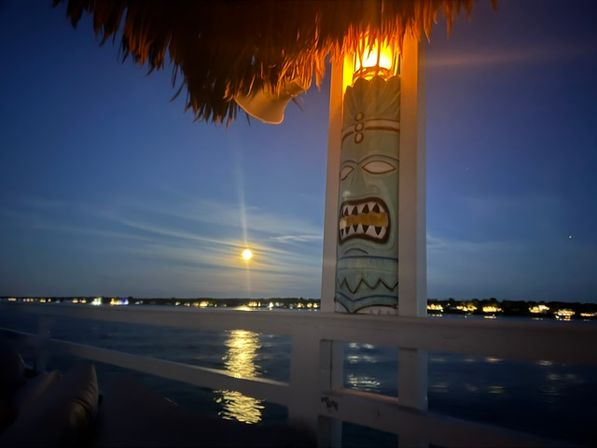 Moonlit waterfront deck with a glowing tiki pole under a thatched roof, full moon reflecting on calm water and twinkling shoreline lights