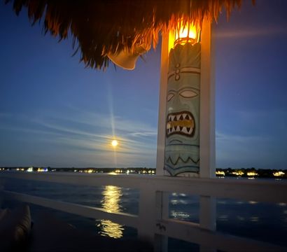 Moonlit waterfront deck with a glowing tiki pole under a thatched roof, full moon reflecting on calm water and twinkling shoreline lights