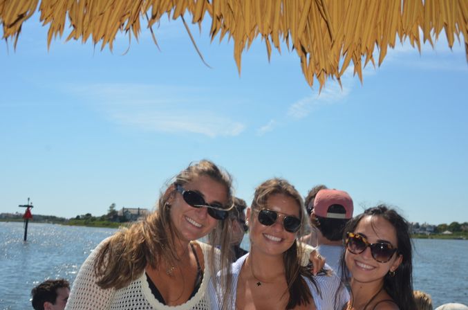 Three smiling friends wearing sunglasses under a straw-thatched umbrella on a sunny waterfront outing with blue sky and a calm bay and shoreline in the background.