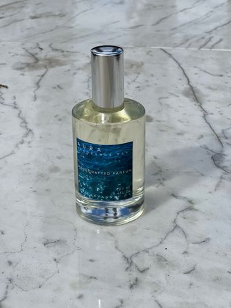 Clear glass perfume bottle with a silver cap and blue ocean-pattern label, standing on a white marble countertop.