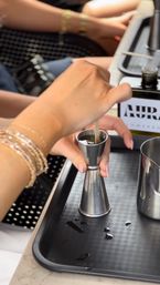 Close-up bartending shot of hands using a stainless-steel jigger to measure and stir a cocktail on a black service tray with small spills and a metal mixing cup nearby.
