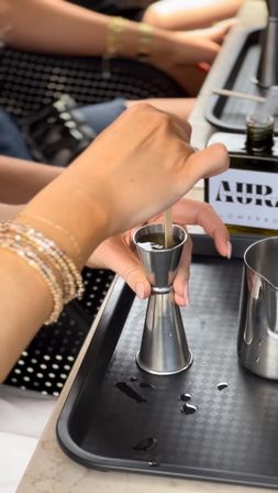 Close-up bartending shot of hands using a stainless-steel jigger to measure and stir a cocktail on a black service tray with small spills and a metal mixing cup nearby.