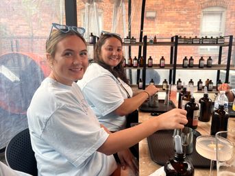 Two smiling women in a tented urban patio mixing cocktails at a communal bar with amber apothecary bottles, metal jiggers and trays, brick building visible outside.
