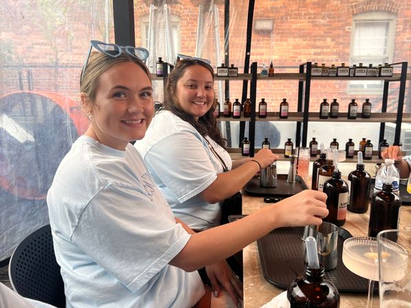 Two smiling women in a tented urban patio mixing cocktails at a communal bar with amber apothecary bottles, metal jiggers and trays, brick building visible outside.