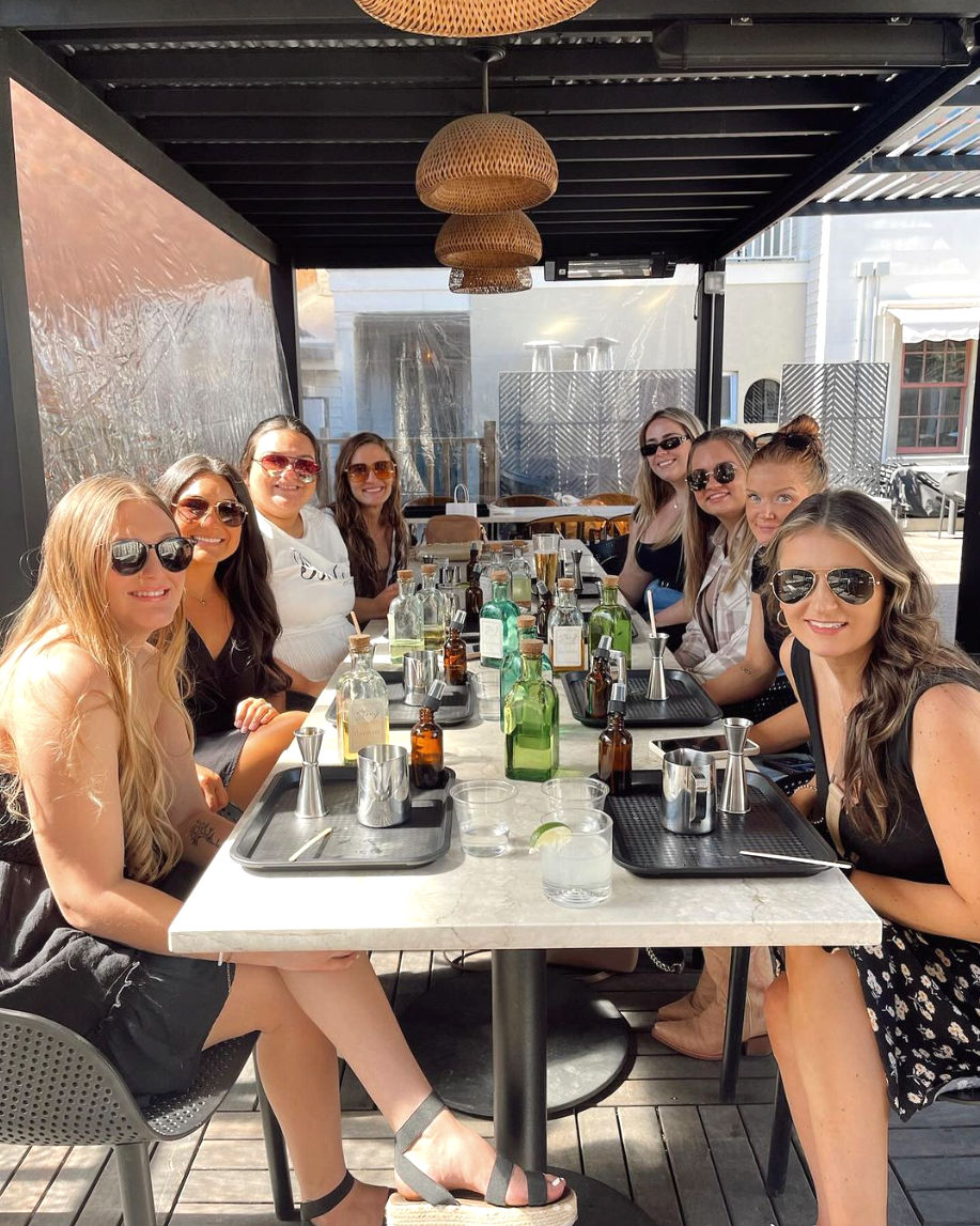 Nine women gathered at a sunny covered outdoor patio table, smiling around a brunch-style cocktail-making setup with bottles, jiggers and wicker pendant lights overhead.