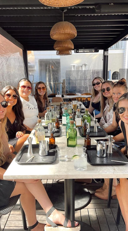 Nine women gathered at a sunny covered outdoor patio table, smiling around a brunch-style cocktail-making setup with bottles, jiggers and wicker pendant lights overhead.