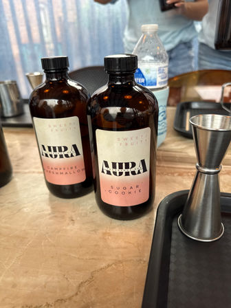 Two amber glass fragrance bottles labeled “Sugar Cookie” and “Campfire Marshmallow” on a marble countertop, with a stainless steel jigger and a water bottle nearby — a cozy, sweet-scented display.