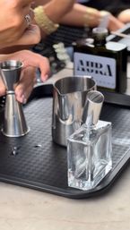 Close-up of a mixology station on a marble countertop: stainless steel jigger and pitcher, clear square bottle with metal pour spout on a black serving tray, and hands preparing a cocktail.