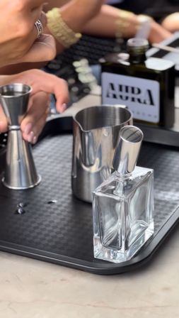 Close-up of a mixology station on a marble countertop: stainless steel jigger and pitcher, clear square bottle with metal pour spout on a black serving tray, and hands preparing a cocktail.