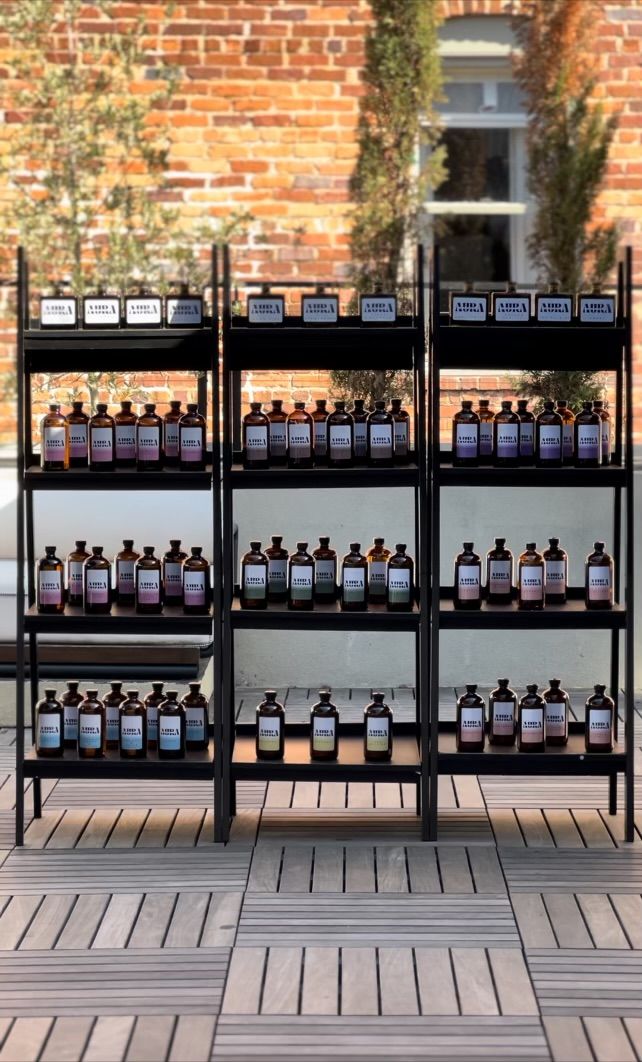 Neat rows of amber apothecary-style bottles with white labels on black metal shelves on a wooden deck, staged as an outdoor patio display against a sunlit red-brick wall with tall potted greenery.