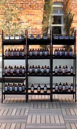 Neat rows of amber apothecary-style bottles with white labels on black metal shelves on a wooden deck, staged as an outdoor patio display against a sunlit red-brick wall with tall potted greenery.