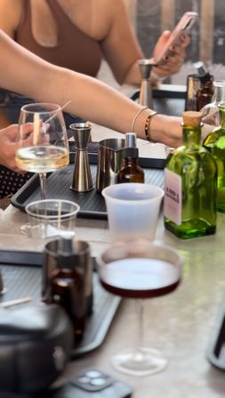 Close-up of a tabletop cocktail-making scene with hands using jiggers and barware on trays, green liquor bottles and amber dropper bottles, a white-wine glass and a dark coupe cocktail, plastic cup and smartphone visible — casual mixology setup.