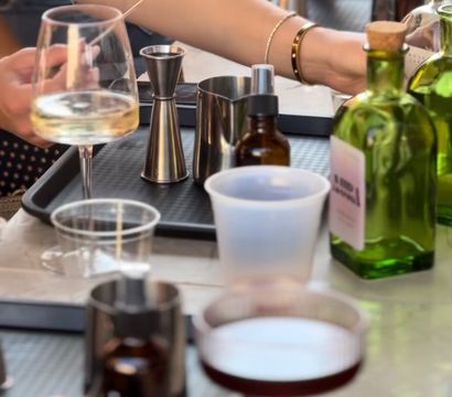 Close-up of a tabletop cocktail-making scene with hands using jiggers and barware on trays, green liquor bottles and amber dropper bottles, a white-wine glass and a dark coupe cocktail, plastic cup and smartphone visible — casual mixology setup.