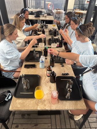 Group of women at a DIY cocktail-making workshop on a covered patio, using droppers and jiggers to fill amber bottles with trays and colorful drinks on a marble table.