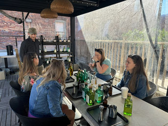 Rooftop patio cocktail tasting with five people gathered around a table of green glass bottles, jiggers and trays, wicker pendant lights overhead and a brick building backdrop.
