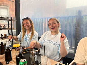 Two smiling young women on a covered city balcony patio mixing drinks at a brunch-style table with bottles, a glass of orange juice, ice and cocktail tools.