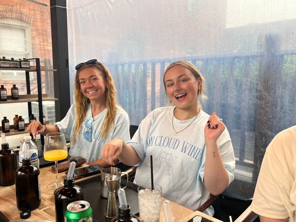 Two smiling young women on a covered city balcony patio mixing drinks at a brunch-style table with bottles, a glass of orange juice, ice and cocktail tools.