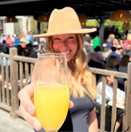 Smiling woman in a tan hat toasting with a mimosa flute on a sunny outdoor brunch patio, blurred crowd and wooden railing in the background