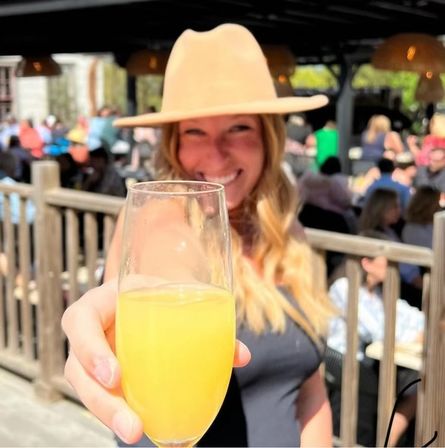 Smiling woman in a tan hat toasting with a mimosa flute on a sunny outdoor brunch patio, blurred crowd and wooden railing in the background