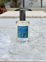 Handcrafted perfume bottle with ocean-blue label and chrome cap sitting on a white marble outdoor patio table, glass reflection and blurred cafe seating in the background