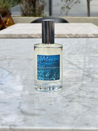 Handcrafted perfume bottle with ocean-blue label and chrome cap sitting on a white marble outdoor patio table, glass reflection and blurred cafe seating in the background